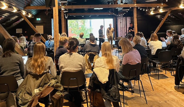 Folk sidder på stole i et lokale og lytter til tre personer, der står foran et stort vindue og taler.